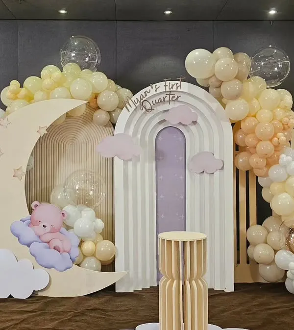 A beautiful celestial-themed setup for "Myan's First Quarter Bash." This decor features a large moon prop with a sleeping teddy bear, a rainbow-arched backdrop, and clouds of soft yellow and white balloons.