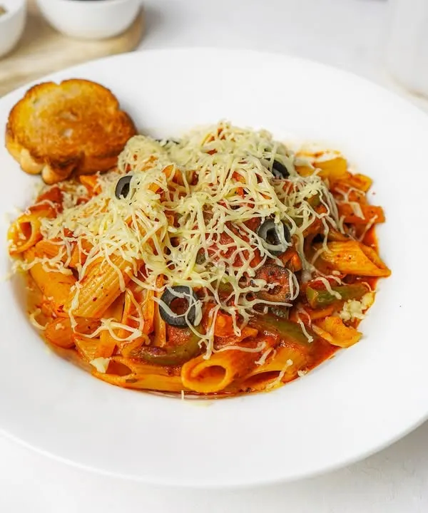 A plate of penne arrabbiata pasta in a rich red sauce, generously topped with shredded cheese and black olives, served with a side of garlic toast. The bright white plate and background make the colors of this Italian dish stand out.