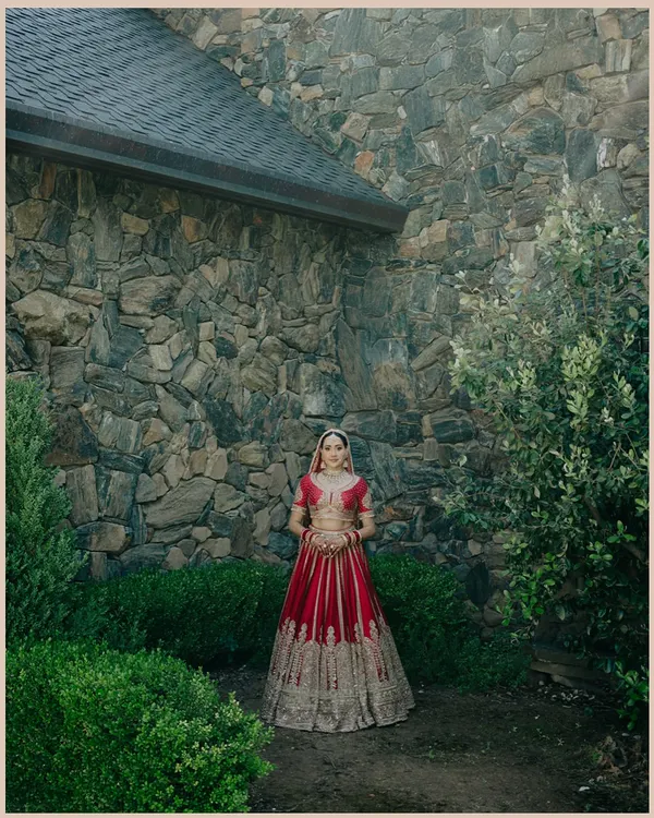 A beautiful portrait of Nita against a rustic stone wall. The contrast between her ornate red lehenga and the natural texture of the wall is striking.