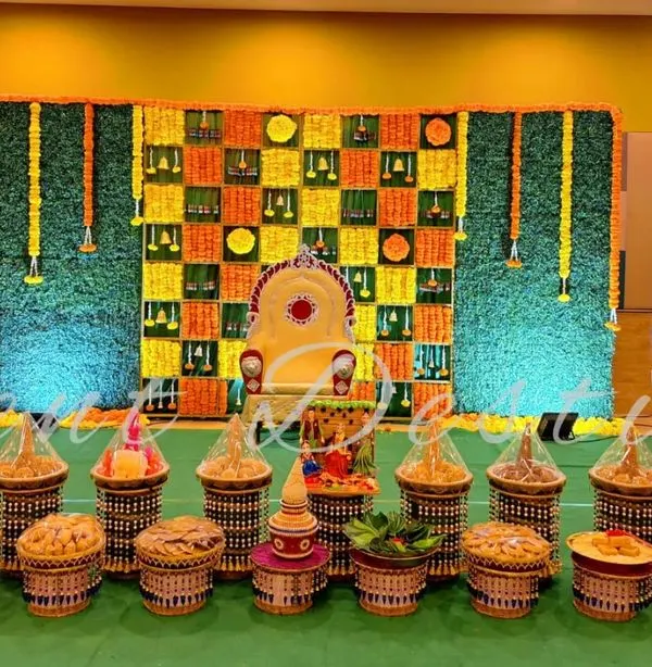 This is a complete setup for a half-saree function, showing both the main stage and the traditional seer plate arrangement. The backdrop features a unique checkerboard pattern of fresh marigolds and green leaves.