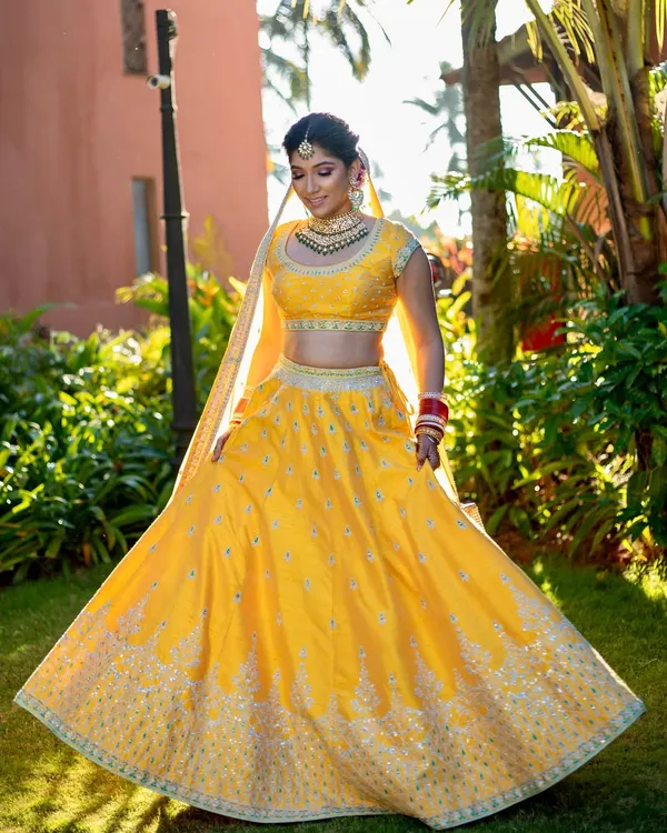 A radiant bride in a sunlit garden. Her makeup is fresh and minimal, with a focus on glowing skin and a soft smile, perfectly complementing her yellow Anita Dongre lehenga.