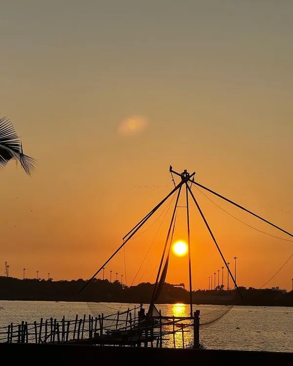 The sun sets behind a Chinese fishing net in Kochi, with a palm tree framing the scene. This is the quintessential Kerala coastal experience, peaceful and picturesque.