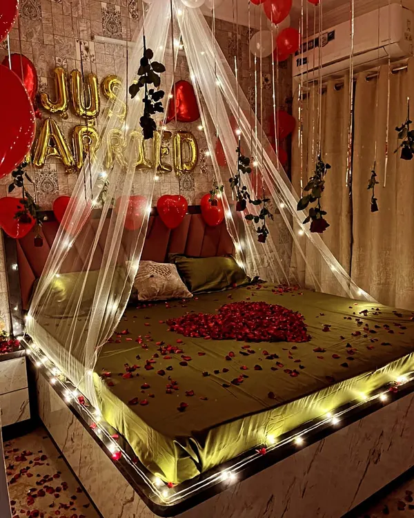 This is my signature 'Just Married' room decoration, perfect for a first night. I create a truly romantic atmosphere using a sheer bed canopy wrapped in warm fairy lights, a heart made of fresh rose petals on the bed, and custom foil balloons. The floor is scattered with petals to complete the immersive experience.