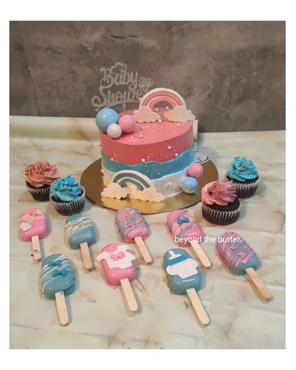 A complete dessert spread for a baby shower. This set includes a beautiful pink and blue rainbow-themed cake, matching cupcakes, and adorable cakesicles shaped like baby clothes.