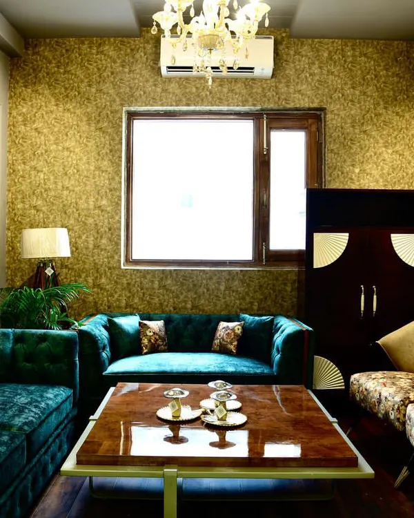 A luxurious living room featuring a deep teal velvet sofa, gold-textured wallpaper, and classic wall moulding. The combination of rich colours and textures creates a warm and inviting atmosphere.