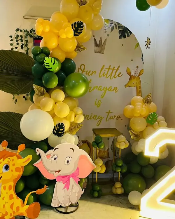 A jungle safari theme for a second birthday. This setup includes a "Wild One" backdrop, cutouts of a giraffe and elephant, a light-up number '2', and a green and yellow balloon garland.