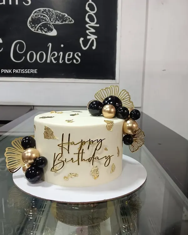 A chic and modern birthday cake with a minimalist aesthetic. The white cake is decorated with black and gold spheres, gold leaf, dried palm fans, and a stylish "Happy Birthday" script topper.