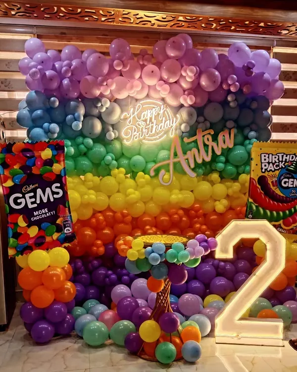 A vibrant and colorful Gems-themed birthday party setup. This large-scale decoration features a full wall of rainbow balloons, a custom neon sign, and giant Cadbury Gems props, perfect for a fun and memorable celebration.