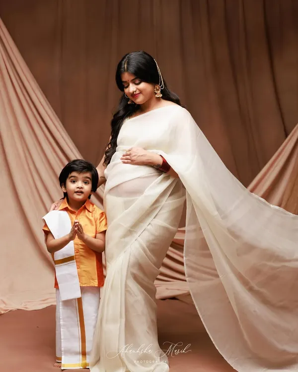 A beautiful maternity portrait celebrating family and tradition. The mother-to-be in a flowing white saree stands with her son, who is dressed in adorable traditional attire.