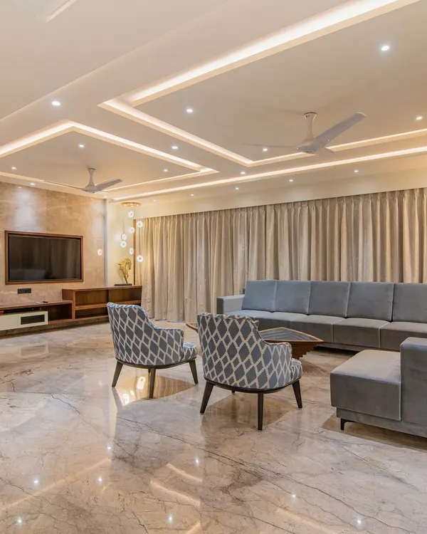 A spacious and bright living room featuring a large sectional sofa, patterned accent chairs, and recessed ceiling lights. The polished marble flooring reflects the light, making the entire area feel open and grand.