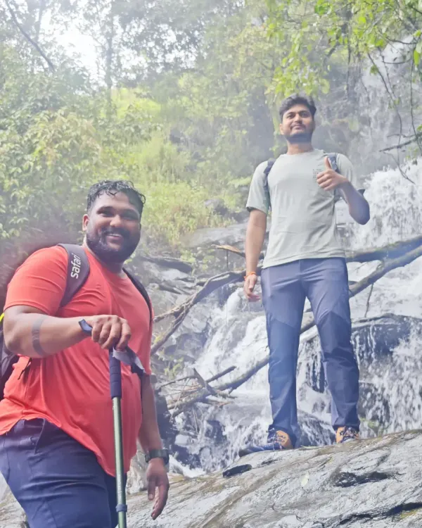 Two trekkers posing in front of a waterfall, fully equipped with backpacks and trekking poles.