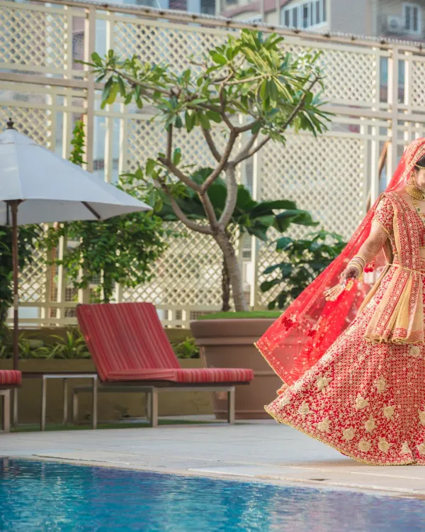 A stunning shot of a bride in a red lehenga walking by the poolside, her veil flowing behind her, creating a sense of movement and grace.