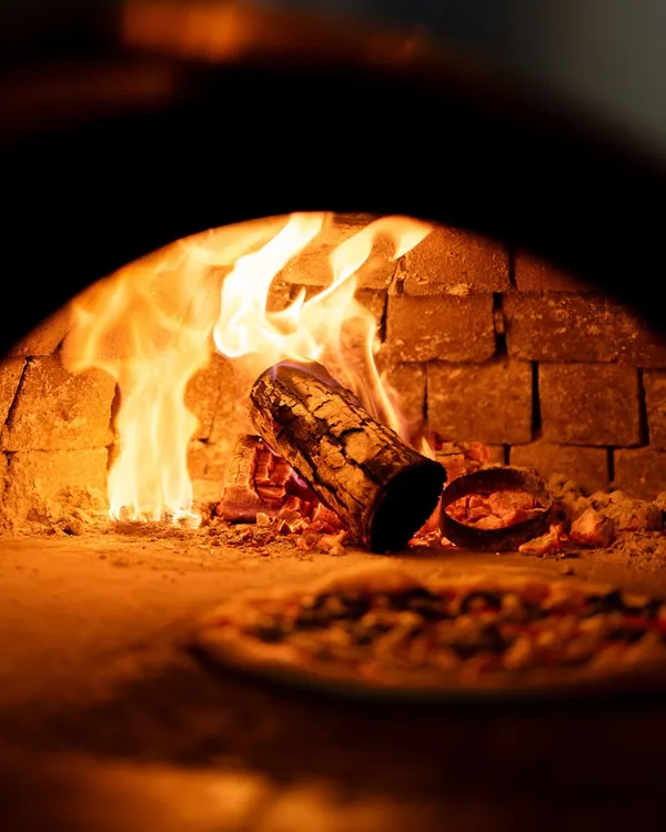 The fire dances just a few steps from your table, wrapping the room in its glow. The crust blisters, the cheese melts, and the air carries you straight to Naples.