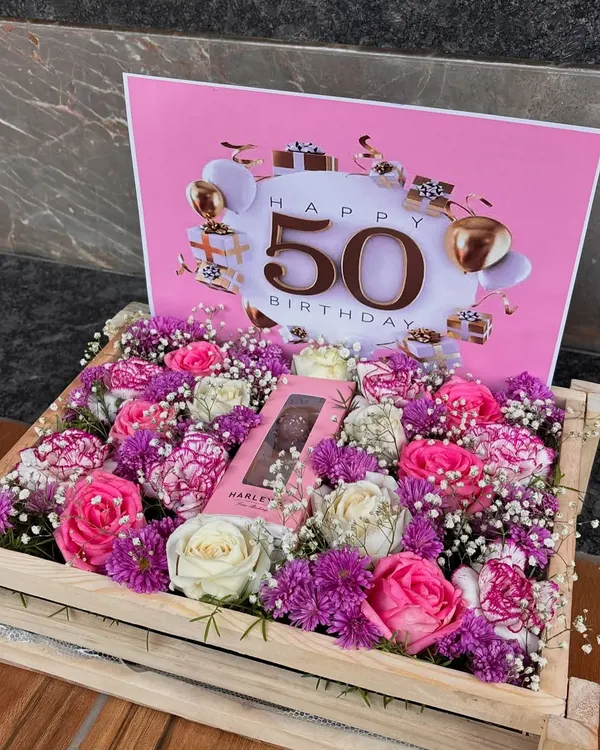 A grand celebration for a milestone. This wooden crate is filled with a vibrant mix of pink and white roses and purple carnations, centered around a chocolate bar and a "Happy 50th Birthday" card.