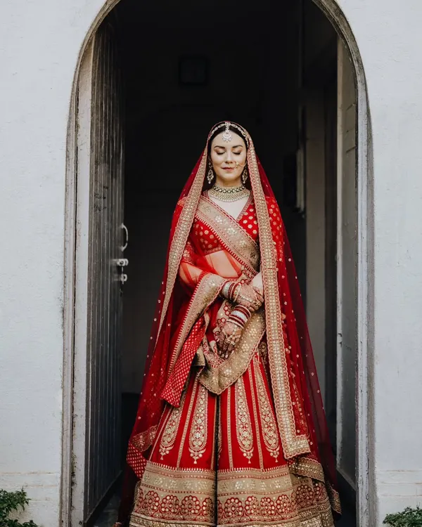 The bride, Rocio, standing in a beautiful archway, looking like a queen in her magnificent red lehenga. A moment of calm elegance before the ceremony.