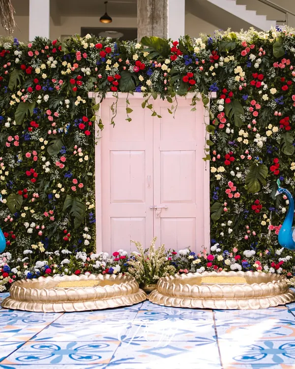 A tropical Haldi dream. We created a stunning backdrop with a wall of fresh green leaves, a pink door, and vibrant florals, complete with peacock and flamingo props for a fun, exotic touch.