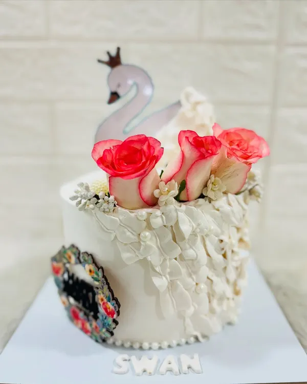 An elegant white cake featuring a graceful swan topper, fresh pink-tipped roses, and delicate lacework-style piping.