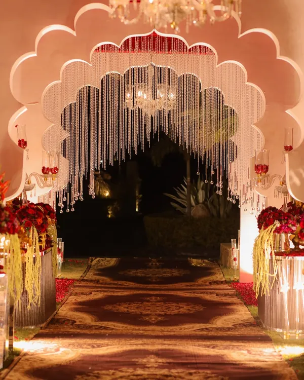 A majestic entrance for a royal-themed wedding, featuring a grand scalloped arch adorned with hanging crystal strings. The path, lined with a traditional patterned carpet and red floral arrangements, exudes opulence.
