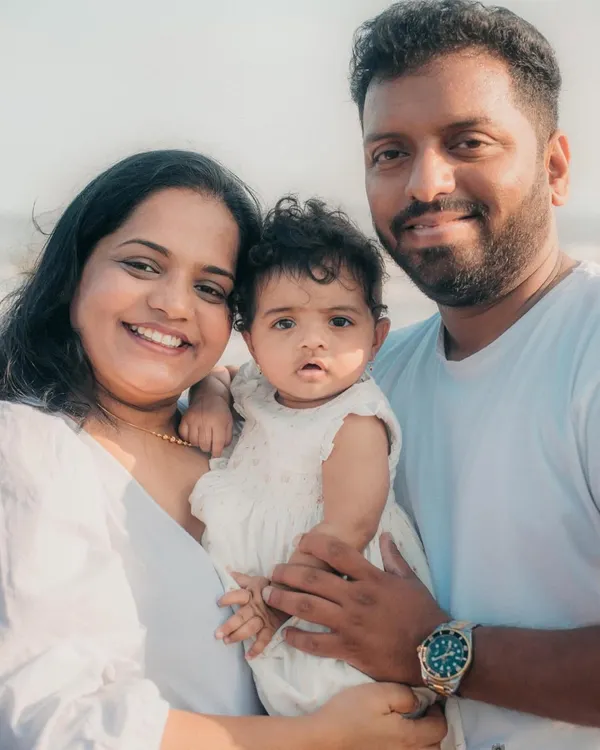 A happy family of three smiling for the camera during their relaxed beach photoshoot. The coordinated white outfits look beautiful against the sea.