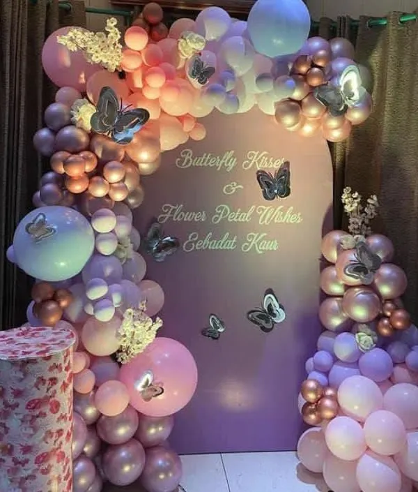 An enchanting butterfly-themed decoration with a purple arched backdrop. The setup is adorned with a beautiful balloon garland in shades of pink and purple, with delicate butterfly accents.