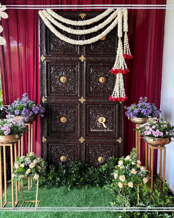 A grand traditional entrance featuring a magnificent carved wooden door, adorned with jasmine garlands. Set against a rich red curtain and surrounded by floral arrangements, this entrance makes a powerful statement.