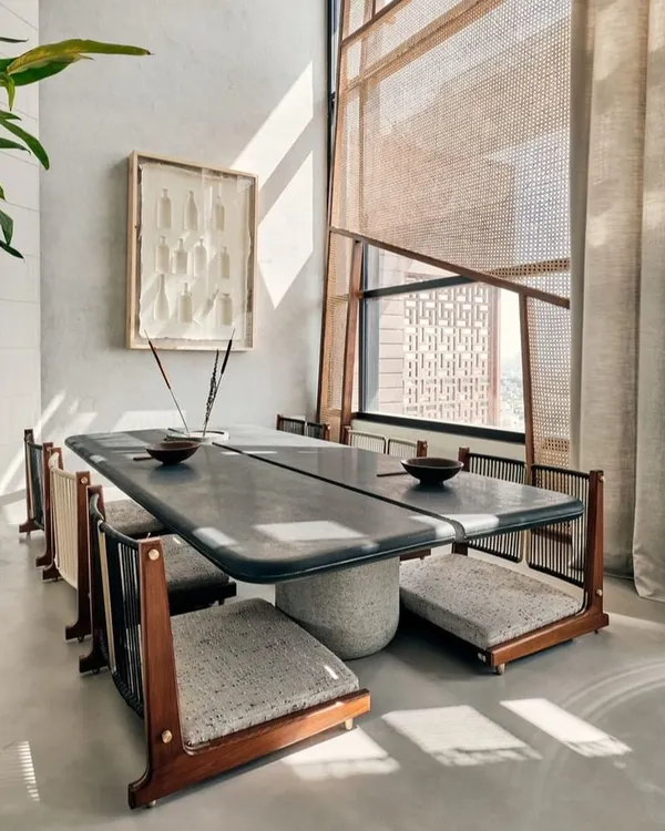 This unique dining setup features low seating and a custom stone-top table, creating a grounded and intimate atmosphere. The design is complemented by natural light filtering through sheer curtains and jali screens.