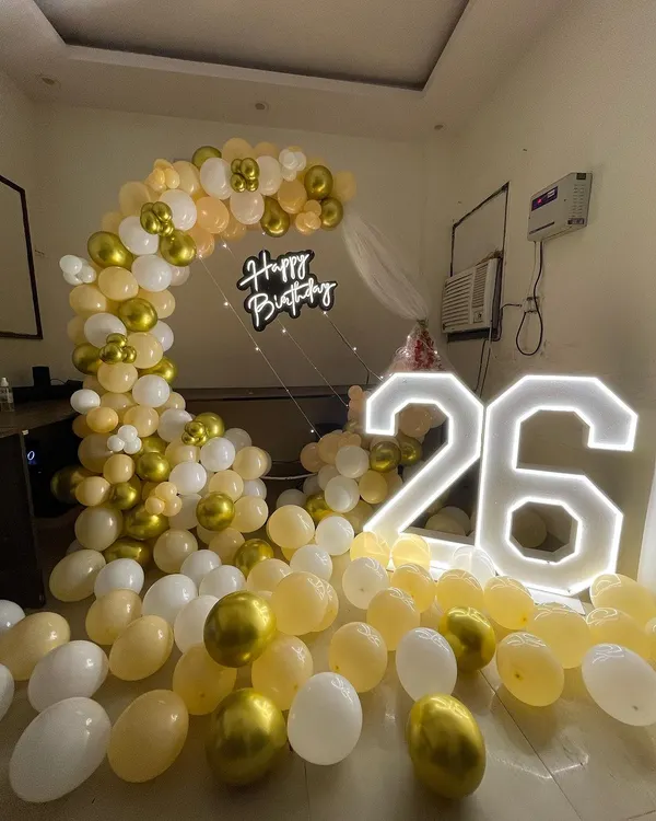 A chic setup for a 26th birthday. This decoration features a half-arch of peach and gold balloons, with large light-up numbers and a "Happy Birthday" neon sign.