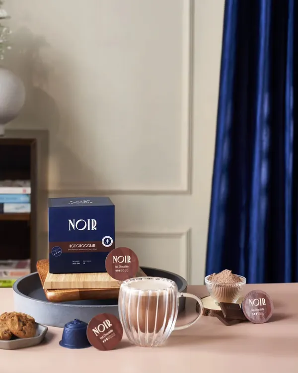 A lifestyle shot for Noir coffee, featuring their hot chocolate pods. The styling with a rich blue curtain and cookies creates a cozy, premium feel for the brand.