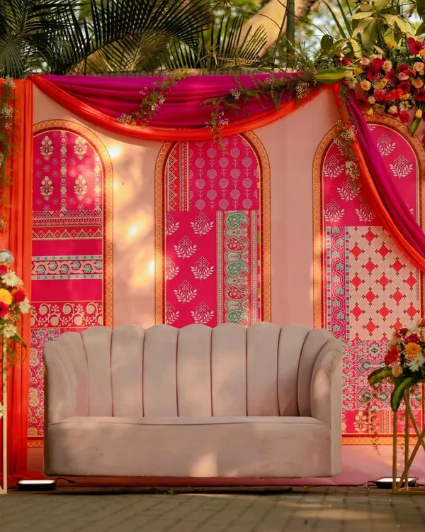 A vibrant Mehendi seating area, featuring a modern scalloped sofa set against a backdrop of colorful block-printed panels and flowing drapes.