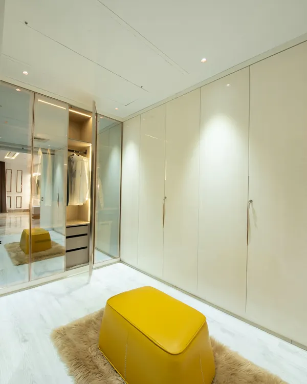 A luxurious walk-in closet concept with high-gloss, floor-to-ceiling wardrobes and a central glass-enclosed island for accessories. A bright yellow ottoman adds a pop of color.