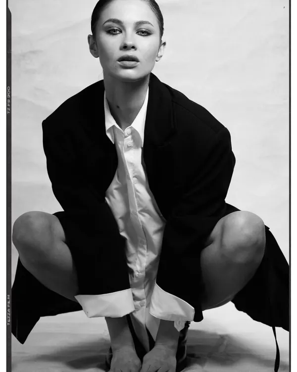 A raw, high-contrast portrait with a minimalist and androgynous feel. The styling consists of an oversized black blazer over a simple white shirt, creating a powerful and confident look with a strong focus on form.