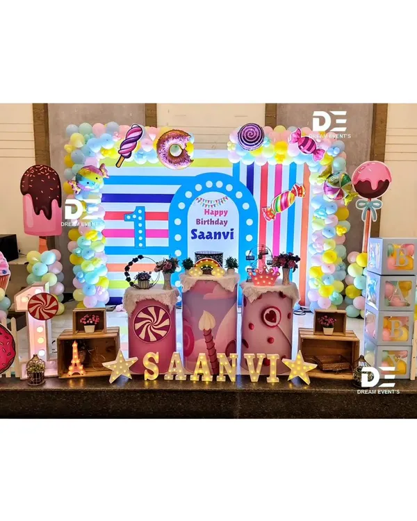 A spectacular Candyland first birthday for Saanvi. This full stage setup is a visual delight with a striped backdrop, giant cupcake and donut props, and a colorful array of balloons.
