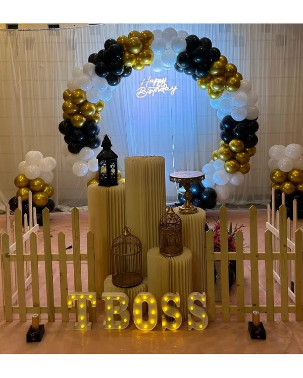 A chic and modern birthday setup with a black, white, and gold color scheme. The decor features a circular balloon arch, a neon sign, and a small picket fence for a stylish touch.