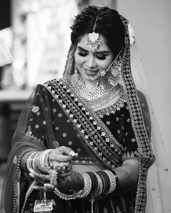 A timeless black and white bridal portrait. This image focuses on the bride's expression as she looks down at her kalire. Converting the photo to monochrome removes distractions, emphasizing the rich textures of her velvet lehenga and the intricate details of her jewelry.