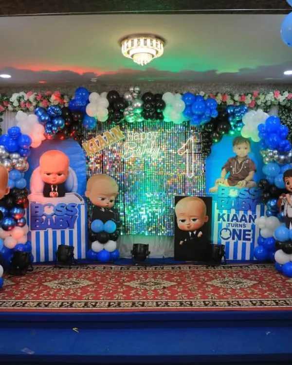A full stage setup for a Boss Baby first birthday party, featuring multiple character cutouts, a silver shimmer backdrop, and themed balloon pillars in a large hall.