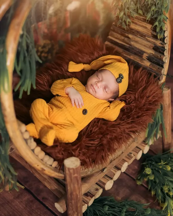 A perfect example of a classic newborn pose. This little one is sleeping peacefully in a rustic, circular wooden prop, dressed in a cozy yellow outfit that contrasts beautifully with the rich, warm tones of the setup.