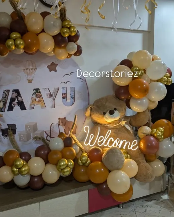 A chic and modern welcome home decor for baby Vaayu. The setup features a custom backdrop, a large plush teddy bear, and a sophisticated balloon arrangement in earthy tones with pops of metallic gold.