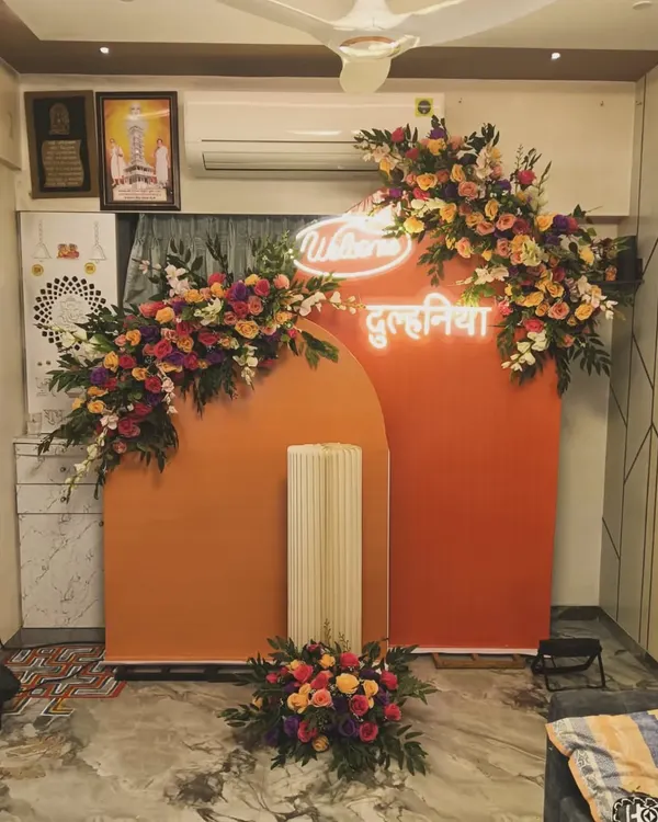 A "Welcome Dulhania" decoration for a new bride's arrival. This home setup features a warm orange backdrop, a custom neon sign, and beautiful floral arrangements.