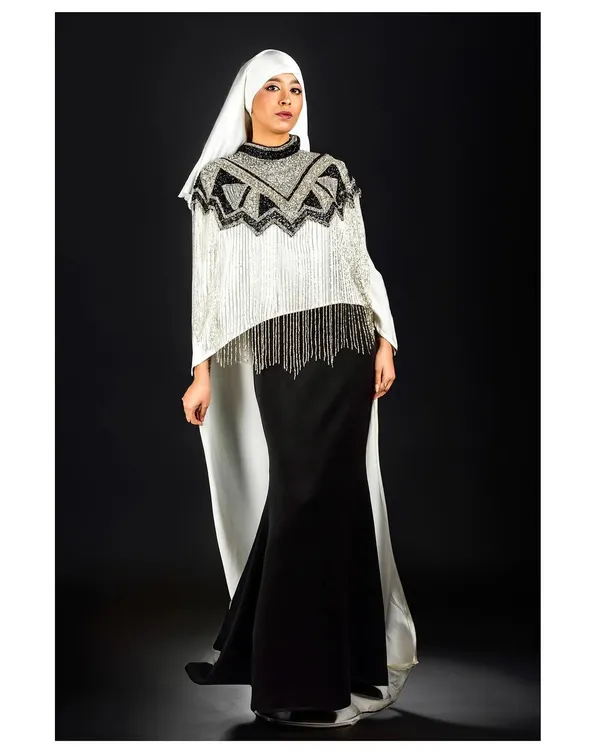 A full-length shot showcasing a designer piece that blends modern structure with flowing elements. The black background makes the white fabric and intricate beadwork pop.