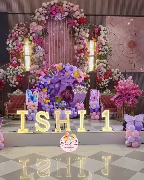 A grand first birthday celebration on a stage, featuring a stunning floral and balloon arch. The setup includes large, illuminated marquee letters spelling 'ISHI 1' and custom butterfly-themed balloon columns.
