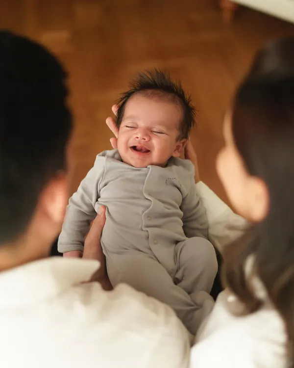 A candid shot of a newborn smiling brightly while being held by their parents, whose faces are softly blurred in the foreground. This over-the-shoulder perspective creates an intimate and heartwarming family moment.