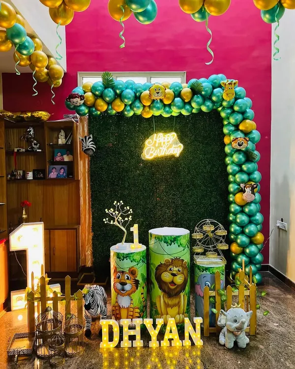 A full view of a jungle safari first birthday party setup in a home. The decoration features a lush green grass wall, a gold and green balloon arch with animal head foils, themed pedestals, and a large light-up number '1'.