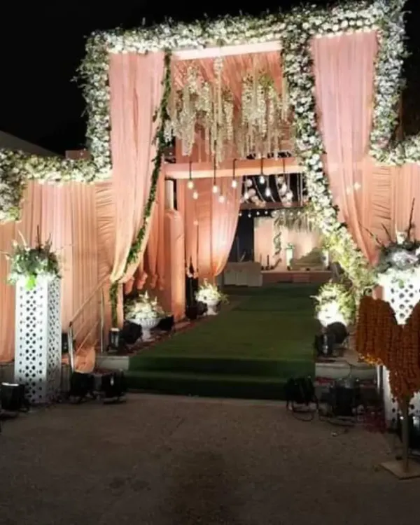A luxurious nighttime wedding entrance featuring soft peach drapes, a full arch of white flowers, and elegant chandeliers. Spotlights illuminate the path, creating a grand and welcoming feel.