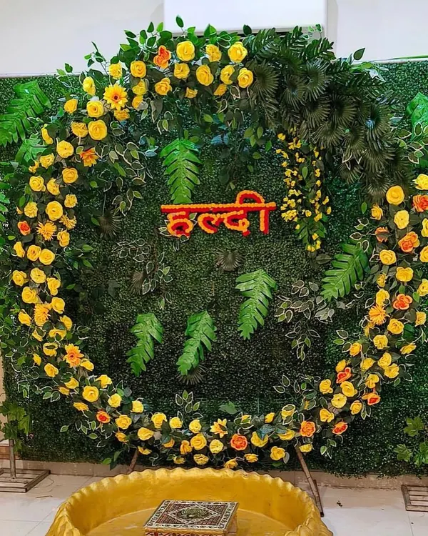 A modern take on Haldi decor, this setup features a circular frame covered in yellow flowers against a dense green foliage wall. The floral 'Haldi' lettering in the center adds a personalized touch to this fresh and stylish photo backdrop.
