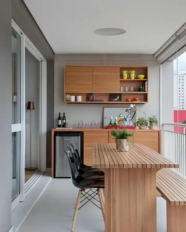 A minimalist and highly functional balcony bar setup. This design features clean wooden cabinetry with open shelving, a built-in mini-fridge, and a matching dining table with a bench. It's a practical solution for creating an al fresco dining experience.