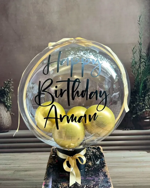 A personalized balloon bouquet featuring a large transparent balloon with a custom 'Happy Birthday' message and smaller gold balloons inside. It's a perfect gift or a centerpiece for a celebration.