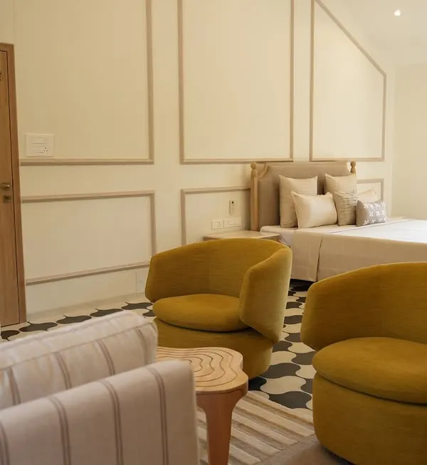 The master bedroom of an Alibaug villa, designed to be a breath of calm. The combination of neutral wall mouldings, a comfortable striped sofa, and bold mustard armchairs creates a space that is effortlessly luxurious.