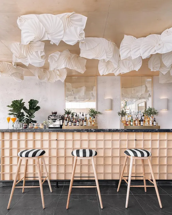 The bar at Brassa features a light wood grid base and bold, black-and-white striped stools. Above, a custom installation of fabric origami lights adds a soft, sculptural element to the ceiling.