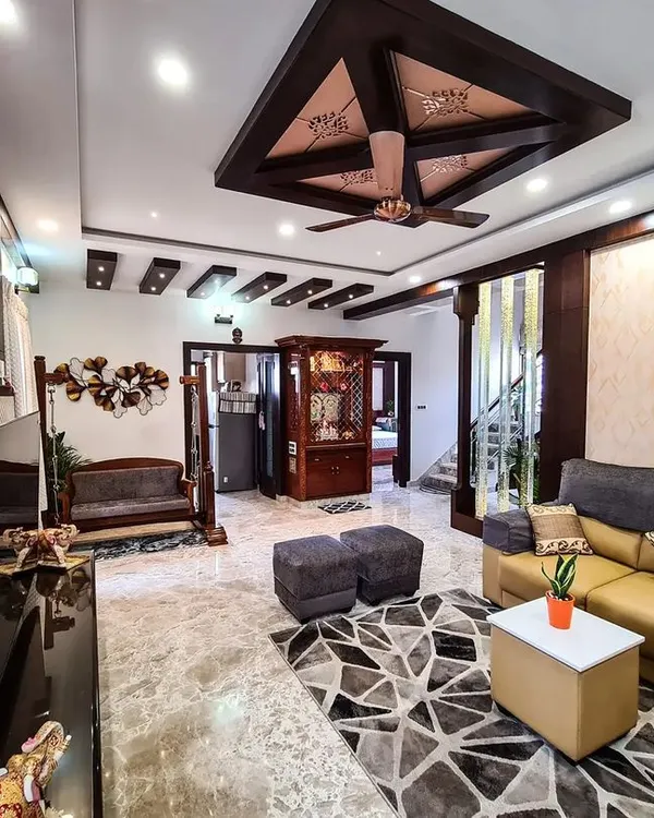 This is a wide view of a family living area where I used marble flooring and a mix of ceiling styles to define the space. The layout includes a comfortable L-shaped sofa, a traditional wooden jhoola, and a glimpse of the ornate pooja mandir, creating a harmonious blend of functions.