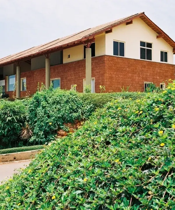 The "Office in a Verandah" in Goa is defined by its large, sheltering roof and laterite base, designed to withstand and celebrate the region's heavy rainfall. The building sits comfortably in its lush, hilly landscape.